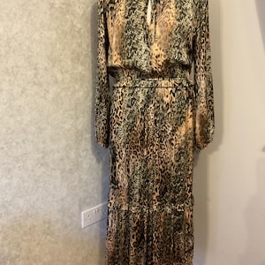 May include: A long, flowing dress with a leopard print design. The dress features long sleeves, a high neckline with a keyhole detail, and a tiered skirt. The color palette includes shades of brown, black, and tan.