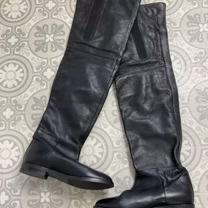 Op de afbeelding: Een paar zwarte leren over-the-knee laarzen. De laarzen hebben een platte zool en een gladde afwerking. De schachten van de laarzen reiken hoog boven de knie. De laarzen worden gepresenteerd op een patroon oppervlak.