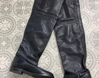 May include: A pair of black leather over-the-knee boots. The boots have a flat sole and a smooth, matte finish. The shaft of the boots extends high above the knee. The boots are displayed on a patterned surface.