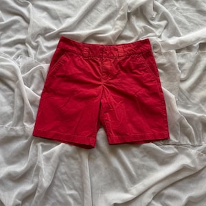 May include: Vibrant red shorts with a classic design. The shorts feature a button and zip fastening, belt loops, and front pockets. The shorts are made of a solid, bright red fabric.