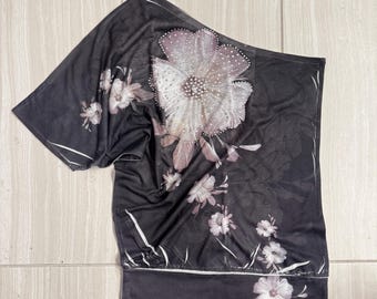 May include: A black one-shoulder top featuring a large, light-colored floral print with scattered silver-toned embellishments on the petals. The garment has a wide, solid black hem at the bottom and a sheer, lightweight fabric construction.