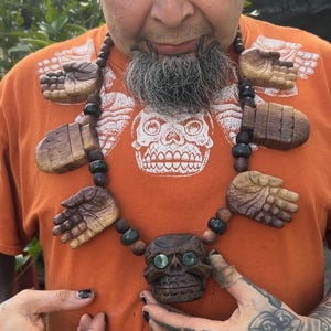 Pode incluir: Um colar de madeira único com pingentes de mãos esculpidas e uma caveira. O colar é feito de madeira marrom escura e é enfiado com contas de madeira. A pessoa na imagem está usando uma camiseta laranja com um gráfico de caveira.
