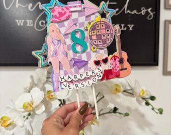 May include: A colorful cake topper with a pop star theme. It features a figure in a sparkly outfit, a disco ball, a guitar, and the number 8. The text "HARPER'S VERSION" is displayed on the bottom. The topper is held by two white sticks.