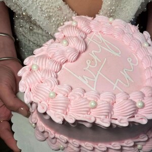 Peut inclure: Un gâteau en forme de cœur rose décoré de glaçage blanc et de pépites de perles. Le gâteau est inscrit avec les mots "Twenty One".