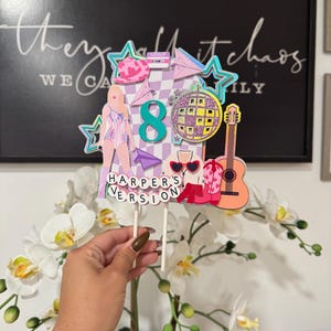 May include: A colorful cake topper with a disco ball, guitar, and the number 8. The topper includes the text "Harper's Version" and a figure in a sparkly outfit. The background features the text "they call it chaos".