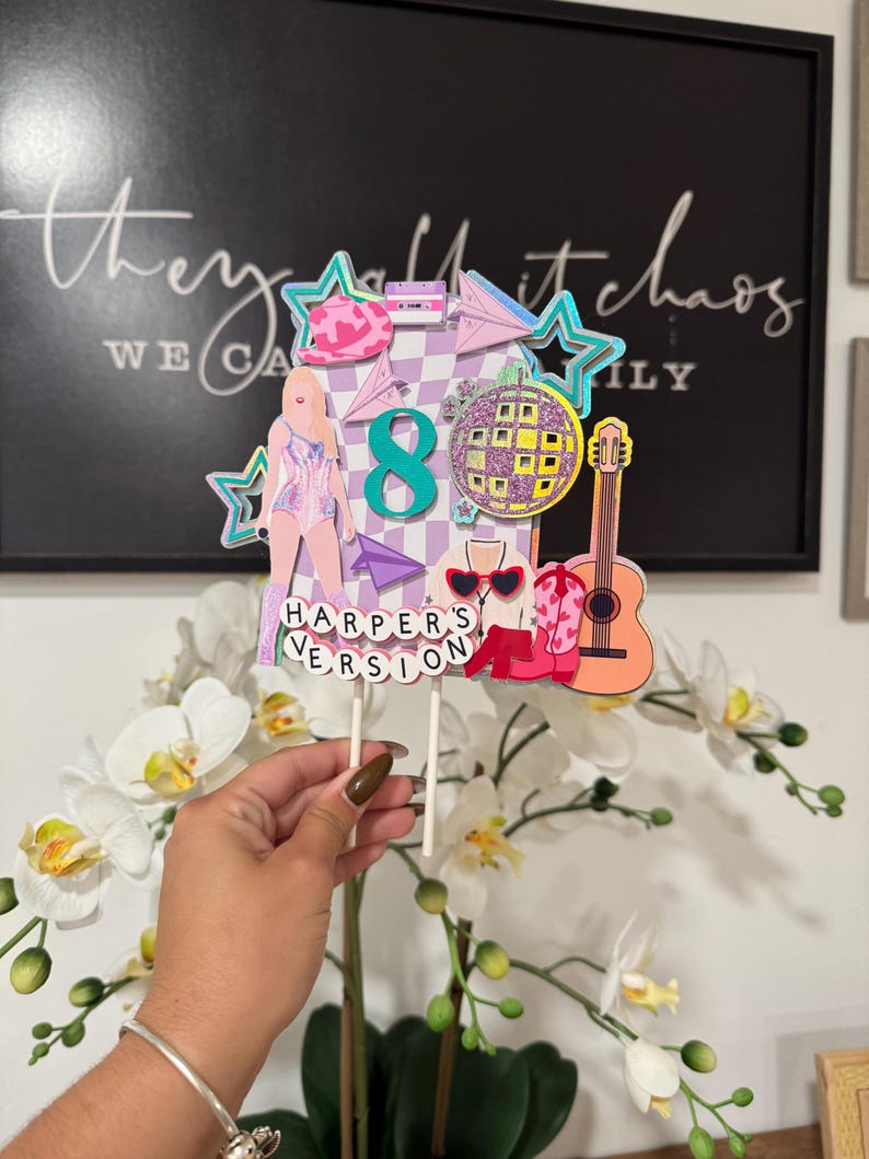 May include: A colorful cake topper with a disco ball, guitar, and the number 8. The topper includes the text "Harper's Version" and a figure in a sparkly outfit. The background features the text "they call it chaos".