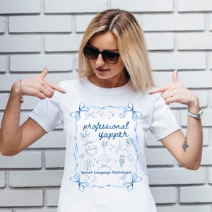 Puede incluir: Camiseta blanca con la frase "professional yapper" en azul, dentro de un marco azul con lazos. El diseño incluye pequeñas ilustraciones azules. El texto "Speech Language Pathologist" está debajo. Una prenda informal para el día a día.