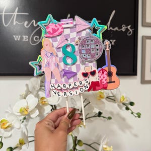 May include: A colorful cake topper with a pop star theme. It features a figure of a singer, a disco ball, a guitar, and the number 8. The text "HARPER'S VERSION" is displayed at the bottom. The topper is held by two white sticks.