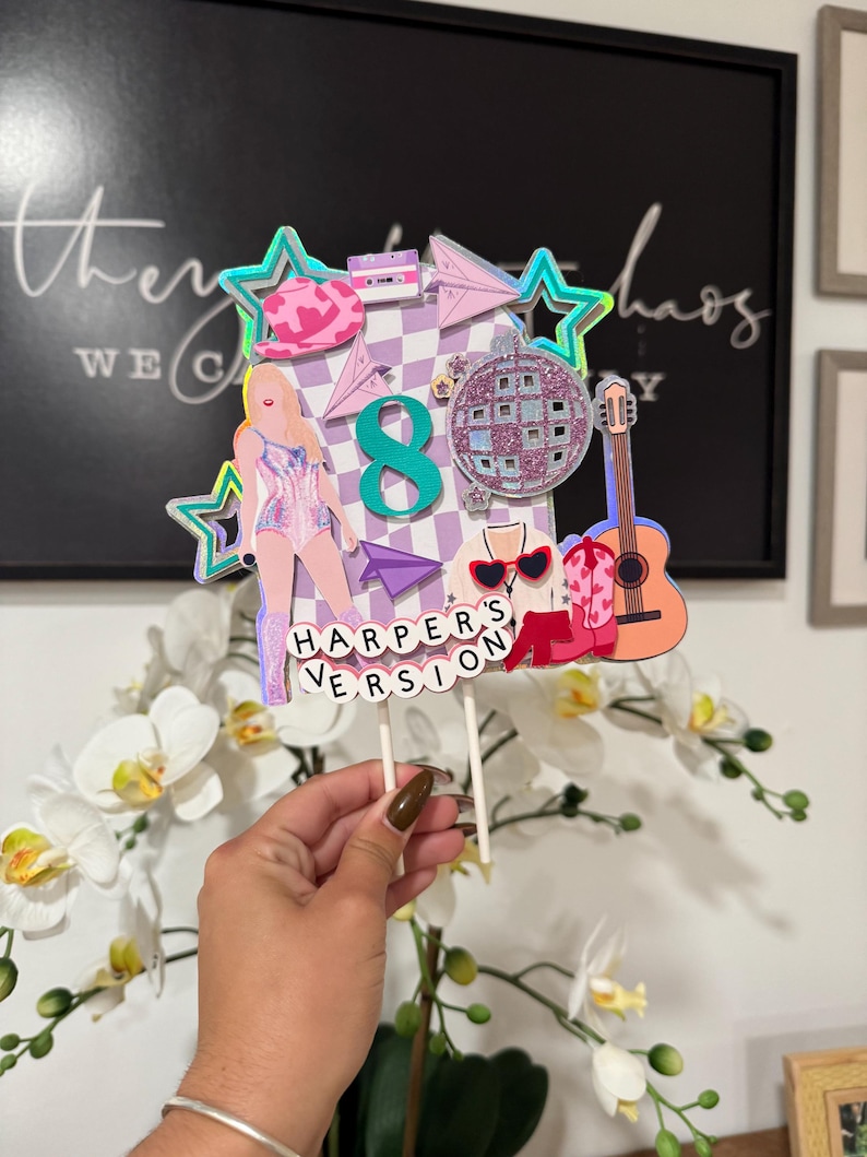 May include: A colorful cake topper with a pop star theme. It features a figure of a singer, a disco ball, a guitar, and the number 8. The text "HARPER'S VERSION" is displayed at the bottom. The topper is held by two white sticks.