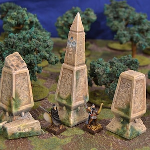 Op de afbeelding: Vier miniatuurstenen obelisken met ingewikkelde gravures en een gezicht op de hoogste. Twee miniatuurfiguren staan voor de obelisken, een met een zwaard en de ander met pijl en boog. De obelisken zijn omringd door groen gebladerte.