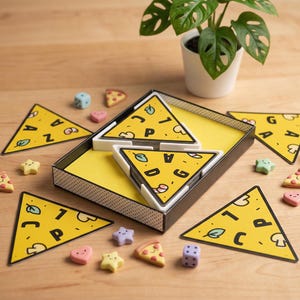 May include: A yellow pizza-themed game with triangular pieces, dice, and small decorative shapes. The pizza slices have letters and food illustrations. The game is set on a wooden surface with a potted plant in the background.