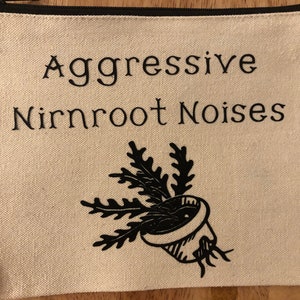 May include: A white canvas zippered pouch with black text that reads "Aggressive Nirnroot Noises" and a black silhouette of a potted plant with roots.