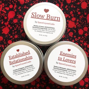Op de afbeelding: Drie zilveren blikken met geurkaarsen met labels met de tekst "Slow Burn", "Established Relationship" en "Enemies to Lovers". De labels bevatten de merknaam "SpaceCoyoteCrafts" en geurbeschrijvingen. De achtergrond is een rood en zwart patroon.