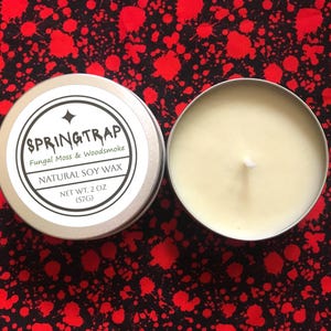 May include: Two silver metal tins of soy wax candles. One tin is open, revealing the candle with a white wick. The other tin has a label that reads "SPRINGTRAP Fungal Moss & Woodsmoke NATURAL SOY WAX NET WT. 2 OZ (57g)". The background is a black fabric with red splatters.