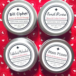 Puede incluir: Cuatro latas plateadas con etiquetas negras y texto blanco. Cada lata contiene velas de cera de soja natural con diferentes aromas: Ámbar y naranja sanguina, Musgo y brasas de cedro, Manzanas y bourbon, y almizcle y vainilla ahumada. Cada lata tiene un peso neto de 2 onzas (57 gramos).