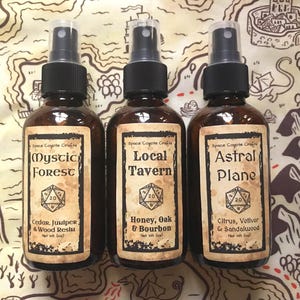 Op de afbeelding: Drie bruine glazen spuitflessen met zwarte sproeikoppen. Elke fles heeft een etiket met de tekst: "Mystic Forest", "Local Tavern" en "Astral Plane". De etiketten bevatten ook geurbeschrijvingen en een dobbelsteenafbeelding. De flessen staan op een stof met een kaartthema.