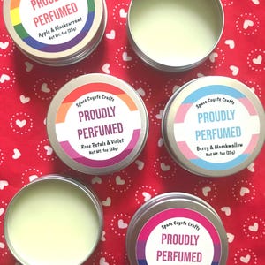 May include: A selection of small, round, silver tins of scented products are displayed on a red background with white hearts. The tins are labelled "PROUDLY PERFUMED" with various scent names and rainbow, orange, blue, and pink accents. Each tin contains 28g of product.