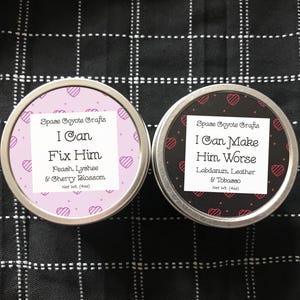 Puede incluir: Dos latas redondas de metal con tapas rosa y negra. La lata rosa tiene una etiqueta blanca que dice "Space Coyote Crafts I Can Fix Him Peach, Lychee & Cherry Blossom Net Wt. (4oz)". La lata negra tiene una etiqueta blanca que dice "Space Coyote Crafts I Can Make Him Worse Labdanum, Leather & Tobacco Net Wt. (4oz)".