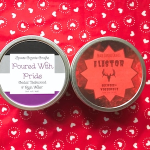May include: Two round metal tins with labels. The tin on the left has a purple, gray, and black label with the text "Space Coyote Crafts Poured With Pride Cedar, Teakwood & Rain Water Net wt 1oz". The tin on the right has a red label with a starburst design and the text "Space Coyote Crafts Alastor Dewood + Voodshote".