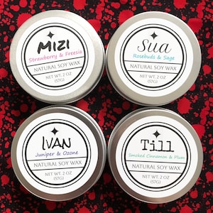 May include: Four silver tins of natural soy wax candles, each with a white label and black border. The labels display the names Mizi, Sua, Ivan, and Till, along with the scent and net weight of 57 grams. The background is red with white splatters.
