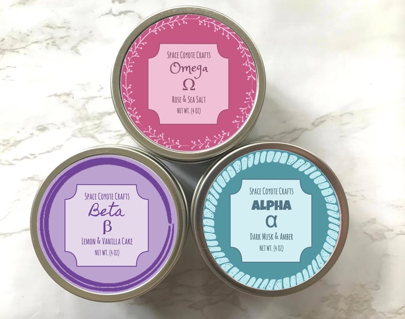 Puede incluir: Tres latas redondas de metal con etiquetas. Las etiquetas son moradas, rosas y azul verdoso. Cada lata tiene una letra griega y un aroma diferentes. La lata morada dice "Beta &beta; Lemon & Vanilla Cake Net Wt. (4 oz)". La lata rosa dice "Omega &Omega; Rose & Sea Salt Net Wt. (4 oz)". La lata azul verdoso dice "Alpha &alpha; Dark Musk & Amber Net Wt. (4 oz)".