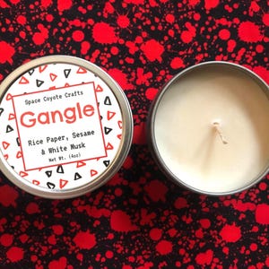 May include: Two round, silver-colored tins of candles. One tin has a label that reads "Gangle" with the scent "Rice Paper, Sesame & White Musk." The other tin is open, revealing a cream-colored candle. Each candle weighs 4 oz.