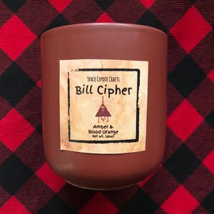 Puede incluir: Un tarro de vela marrón con una etiqueta que dice "SPACE COYOTE CRAFTS Bill Cipher Amber & Blood Orange Net Wt. (8 oz)".