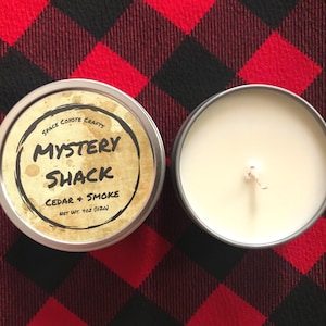 Puede incluir: Dos velas de lata plateadas sobre una superficie a cuadros rojos y negros. Una vela tiene una tapa con el texto "Mystery Shack Cedar + Smoke" y "Space Coyote Crafts". La otra vela está abierta, revelando un interior de cera blanca.