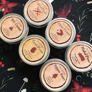 Puede incluir: Colección de siete latas redondas de metal con etiquetas de "Space Coyote Crafts". Cada lata tiene un nombre y aroma diferente, incluyendo "Bill Cipher", "Ford Pines" y "Dipper Pines". Las etiquetas son marrones con ilustraciones.