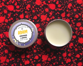 May include: Two silver tins of balm on a red and black patterned background. One tin is open, showing a light yellow balm. The closed tin has a label with the text "ADAM AMBERWOOD & JUNIPER Net Wt. 1 oz (28g)".