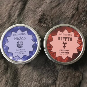 May include: Two round metal tins with labels. The tin on the left is blue with stars and the text "SPACE COYOTE CRAFTS Stolas Rain Water & Sherry Blossom Net wt. (4oz)". The tin on the right is red with flames and the text "SPACE COYOTE CRAFTS BLITZO CINNAMON + WOODSMOKE Net Wt. (4oz)".