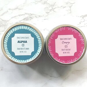 Puede incluir: Dos peque&ntilde;as latas redondas de metal con etiquetas. La lata de la izquierda es azul con letras blancas y dice "Space Coyote Crafts Alpha &alpha; Dark Musk & Amber Net Wt. (4 oz)". La lata de la derecha es rosa con letras blancas y dice "Space Coyote Crafts Omega &Omega; Rose & Sea Salt Net Wt. (4 oz)".