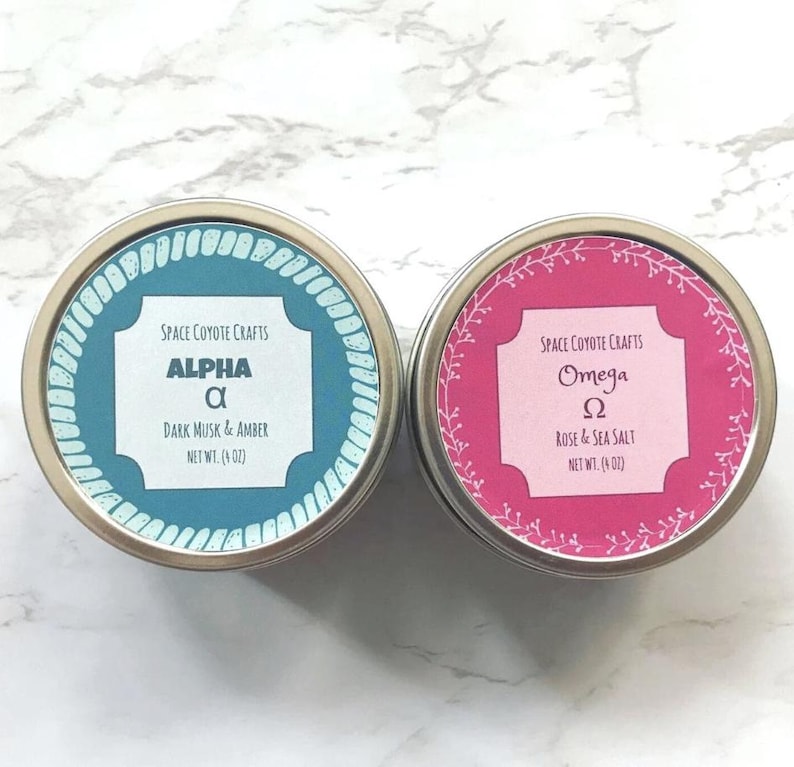 Puede incluir: Dos peque&ntilde;as latas redondas de metal con etiquetas. La lata de la izquierda es azul con letras blancas y dice "Space Coyote Crafts Alpha &alpha; Dark Musk & Amber Net Wt. (4 oz)". La lata de la derecha es rosa con letras blancas y dice "Space Coyote Crafts Omega &Omega; Rose & Sea Salt Net Wt. (4 oz)".