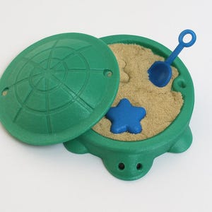 May include: A green turtle-shaped sandbox with a lid, filled with sand, a blue star, and a blue shovel. The lid is partially open, revealing the sand and accessories. The sandbox is designed for children's outdoor play.