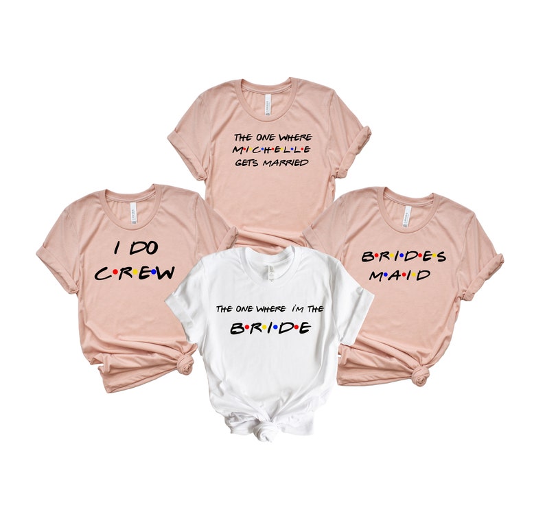 May include: Four t-shirts, three in a light pink and one in white. The shirts have text in a colorful font. The text on the pink shirts reads: "THE ONE WHERE MICHELLE GETS MARRIED", "I DO CREW", and "BRIDES MAID". The white shirt says "THE ONE WHERE I'M THE BRIDE".