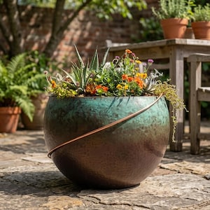 May include: A round, hammered metal planter with a gradient finish transitioning from a dark brown base to a vibrant teal top. A thin, curved copper-colored metal rod arches across the front. The planter is filled with various green succulents and small orange, yellow, and purple flowers.