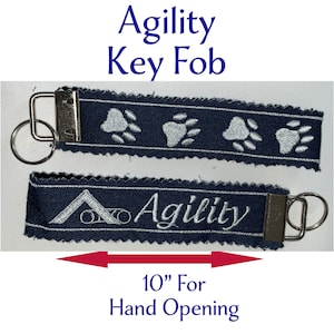 May include: Two navy blue key fobs with silver hardware. One fob has embroidered paw prints, the other has the word "Agility" and a dog agility symbol. The text "Agility Key Fob" and "10" For Hand Opening" are also visible.
