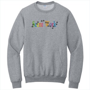 May include: A heather gray crewneck sweatshirt featuring the embroidered text Scent Work in multicolored letters, surrounded by small black paw print icons. The garment includes a front kangaroo pocket and long sleeves with ribbed cuffs and hem.