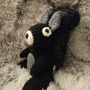 May include: Black and gray crocheted squirrel plush toy with white eyes. The toy is sitting on a fluffy gray surface.