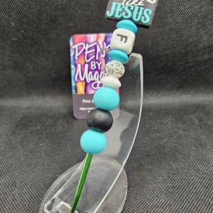 May include: A beaded pen with a green stem and a decorative top. The top features a square with the words "LOVE like JESUS" and various beads in teal, white, black, and silver. A business card is visible in the background.