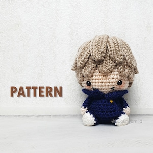 May include: Crocheted amigurumi doll with brown hair, wearing a blue sweater and white shoes. The doll is sitting on a white surface. The text "PATTERN" is visible in the background.