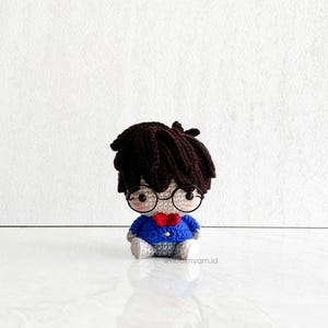 May include: A crocheted plush toy of a person wearing round glasses, a blue sweater, and a red bow tie. The toy has brown hair and a light brown face.