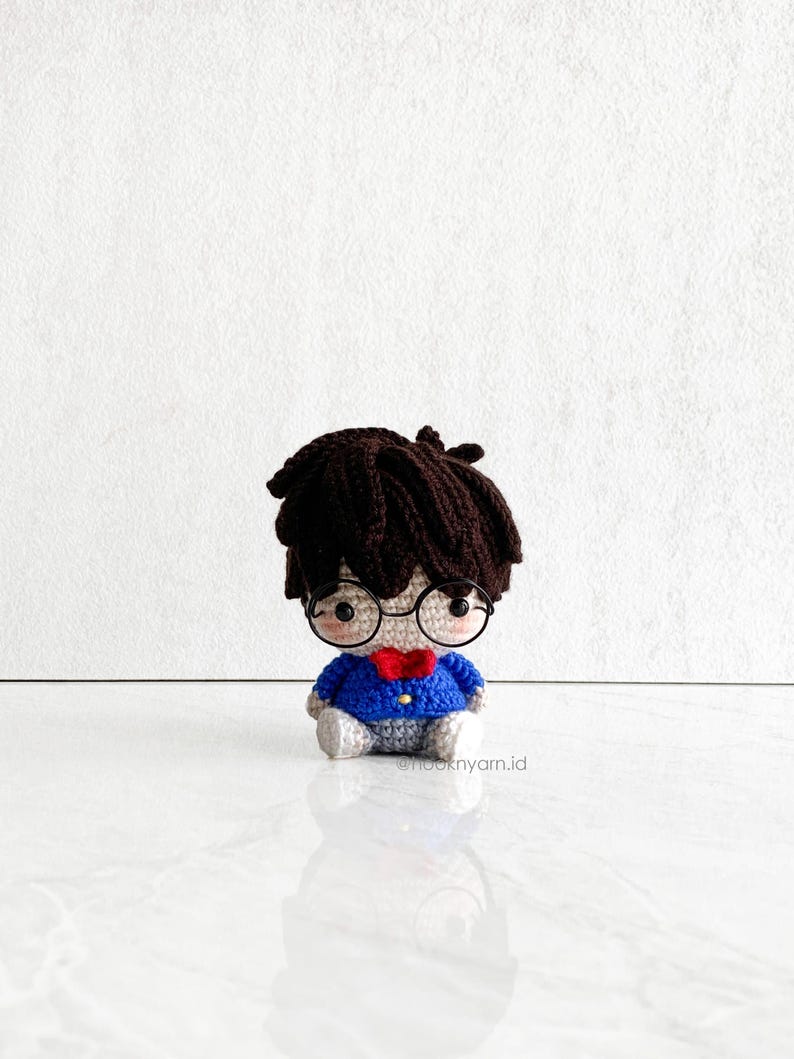 May include: A crocheted plush toy of a person wearing round glasses, a blue sweater, and a red bow tie. The toy has brown hair and a light brown face.