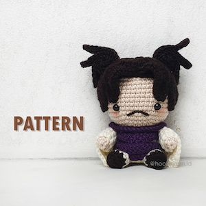 May include: Crocheted amigurumi doll with brown hair in pigtails, wearing a purple shirt and white jacket. The doll has a sad expression and is sitting on a white surface. The text "PATTERN" is visible in the image.