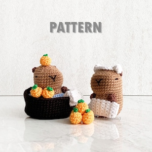 Op de afbeelding: Twee gehaakte bruine capybara knuffels, een in een bruin bad met oranje gehaakte vruchten, de andere houdt een witte handdoek vast. De bijschrift luidt "PATTERN".