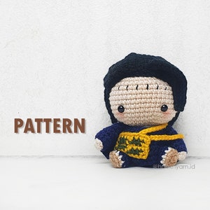 May include: Crocheted amigurumi doll with black hair, a blue and yellow outfit, and a cute expression. The doll is sitting on a white surface. The text "PATTERN" is visible in the image.