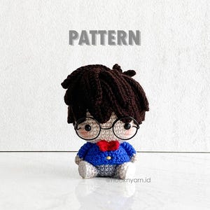 May include: Crocheted amigurumi doll with brown hair, wearing blue and grey clothing and round glasses. The doll is sitting on a white surface. The text "PATTERN" is above the doll.