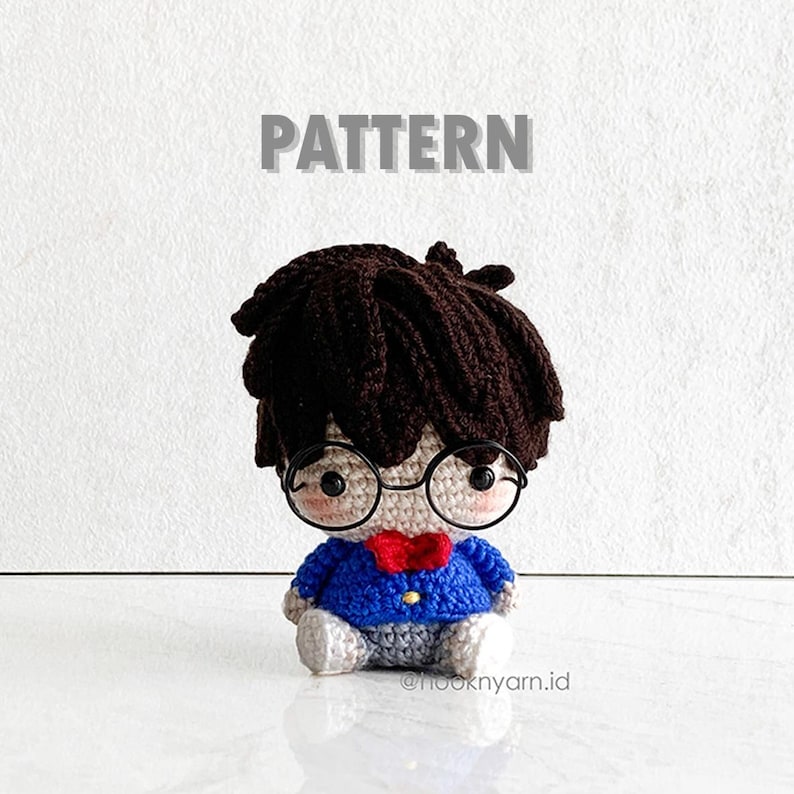 May include: Crocheted amigurumi doll with brown hair, wearing blue and grey clothing and round glasses. The doll is sitting on a white surface. The text "PATTERN" is above the doll.