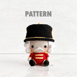 May include: A crocheted toy nutcracker with a black hat, white beard, and red and gold uniform. The toy is sitting on a white surface. The text "PATTERN" is above the toy and the text "@hooknyarn.id" is below the toy.