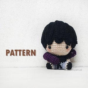 May include: Crocheted amigurumi doll with black hair, purple sleeves, and blue trousers. The doll is sitting on a white surface. The text "PATTERN" is visible in the image.